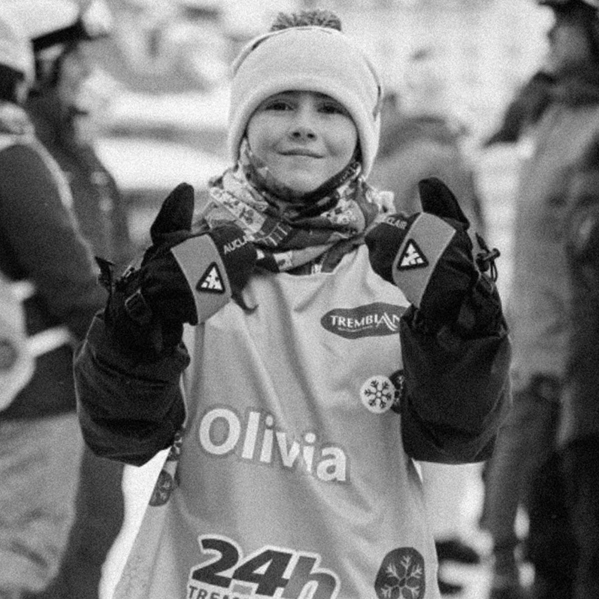 A black and white photo of a woman in winter gear, including a beanie, gloves, and a bib labeled 'Olivia' and 'Tremplin', posing with her gloved hands raised.