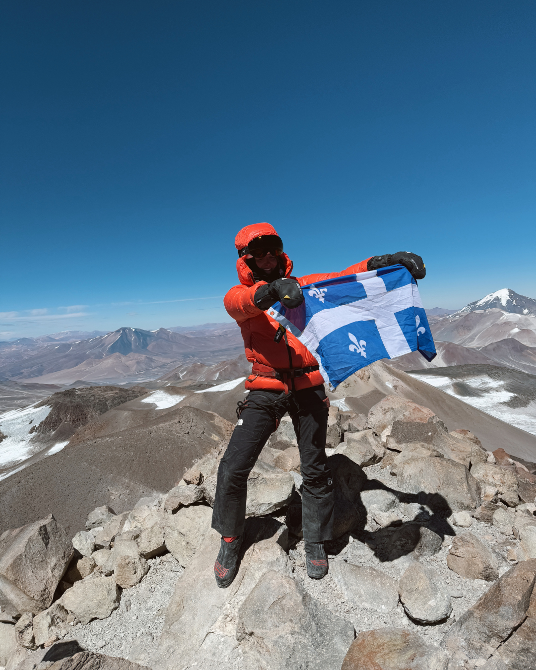 Person on a mountain top holding a flag, with a clear blue sky and mountainous landscape in the background.