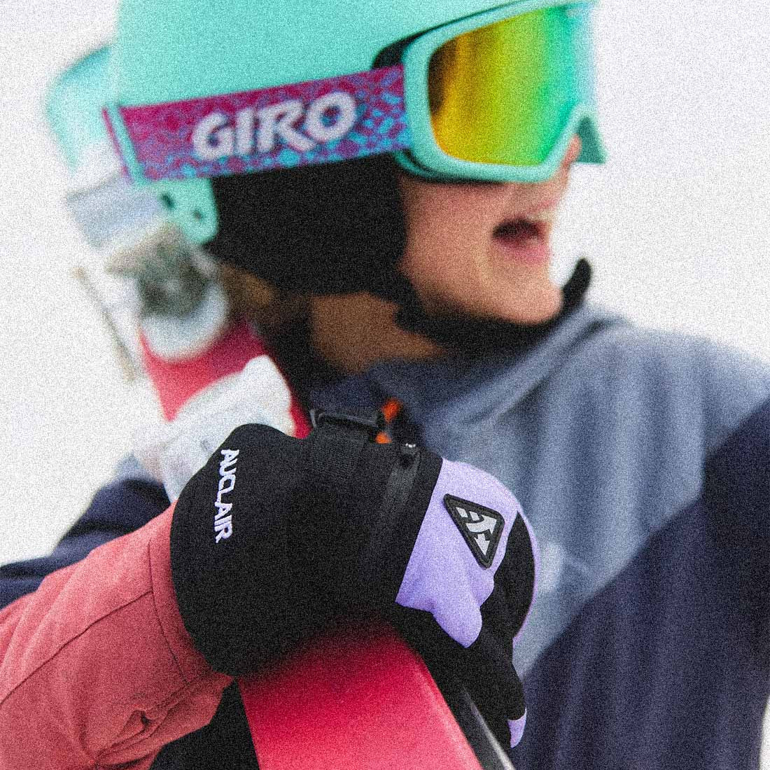 A close-up lifestyle shot of a skier wearing a mint green Giro helmet and mirrored ski goggles, holding a pink ski. The skier is wearing black and lavender gloves with visible 'ANDUR' branding.