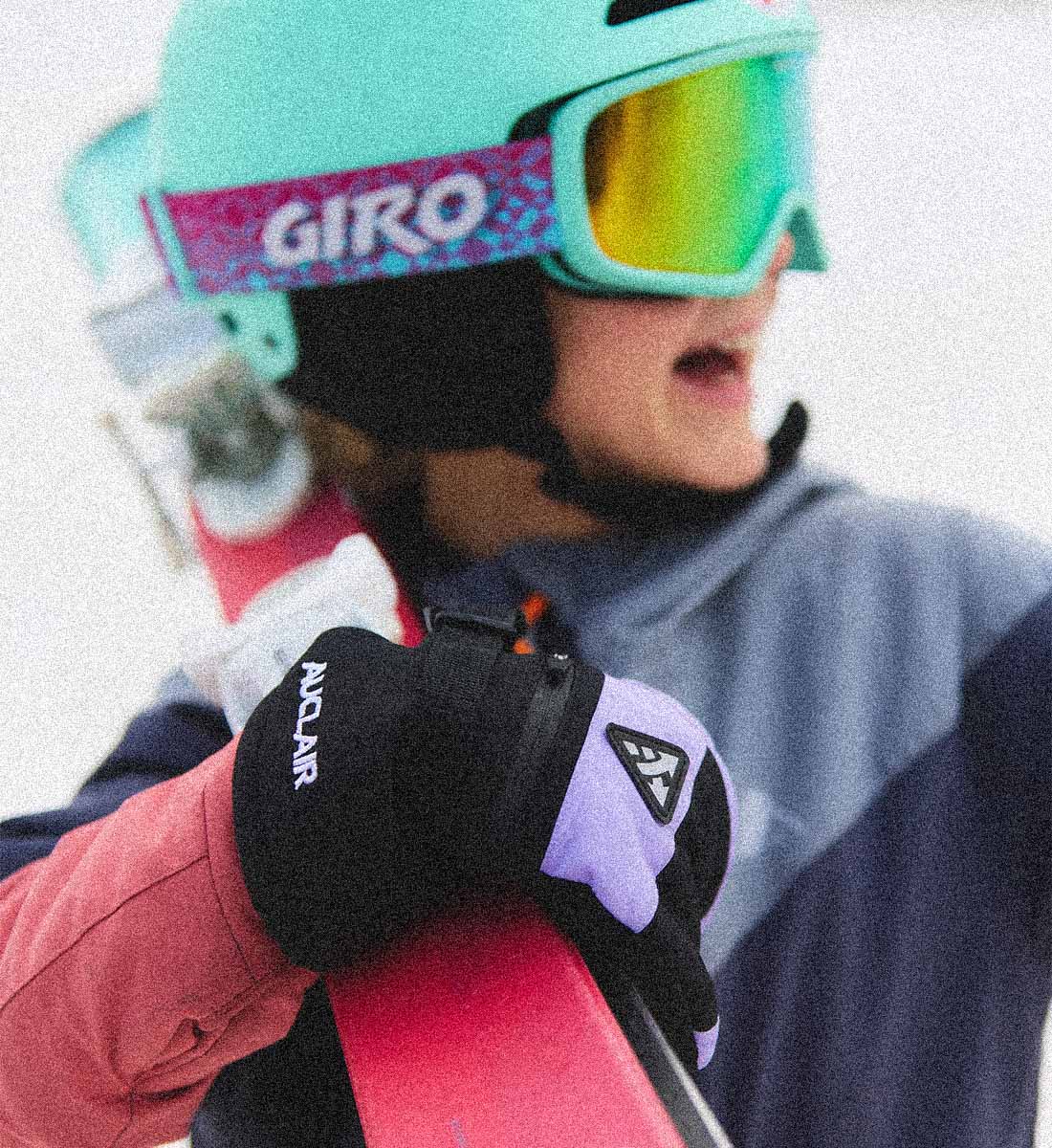 A close-up lifestyle shot of a skier wearing a mint green Giro helmet and mirrored ski goggles, holding a pink ski. The skier is wearing black and lavender gloves with visible 'ANDUR' branding.