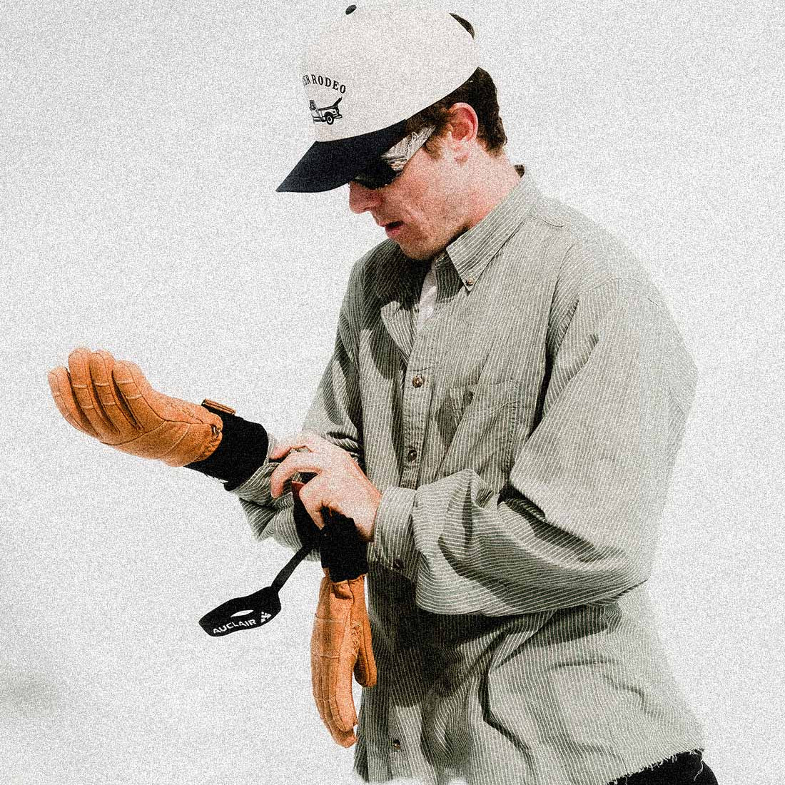 A man wearing a light gray button-down shirt, a white and black cap, and sunglasses is shown from the waist up, adjusting a tan leather glove on his wrist while holding another glove and a black accessory against a textured off-white background.