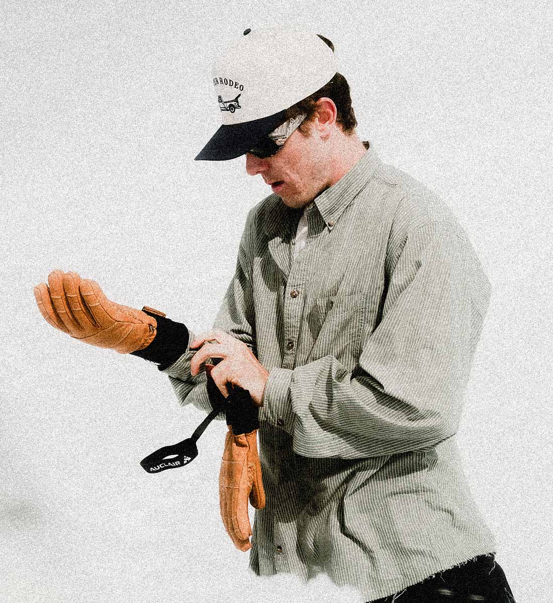 A man wearing a light gray button-down shirt, a white and black cap, and sunglasses is shown from the waist up, adjusting a tan leather glove on his wrist while holding another glove and a black accessory against a textured off-white background.