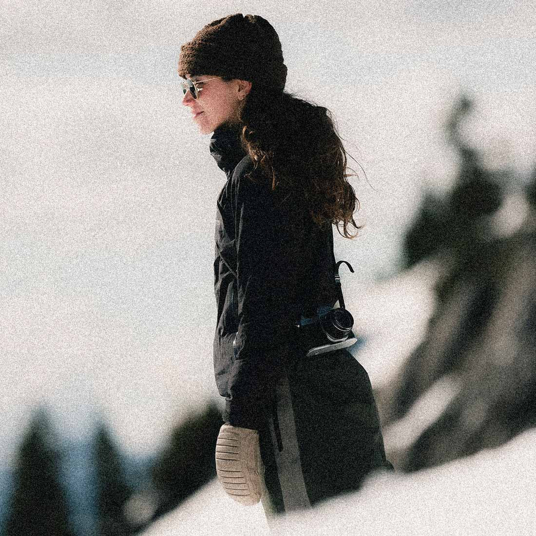 A woman in a black winter jacket, matching beanie, and knit mitts stands in a snowy outdoor setting, looking off-camera. The background is heavily blurred with a grainy, stylized filter.
