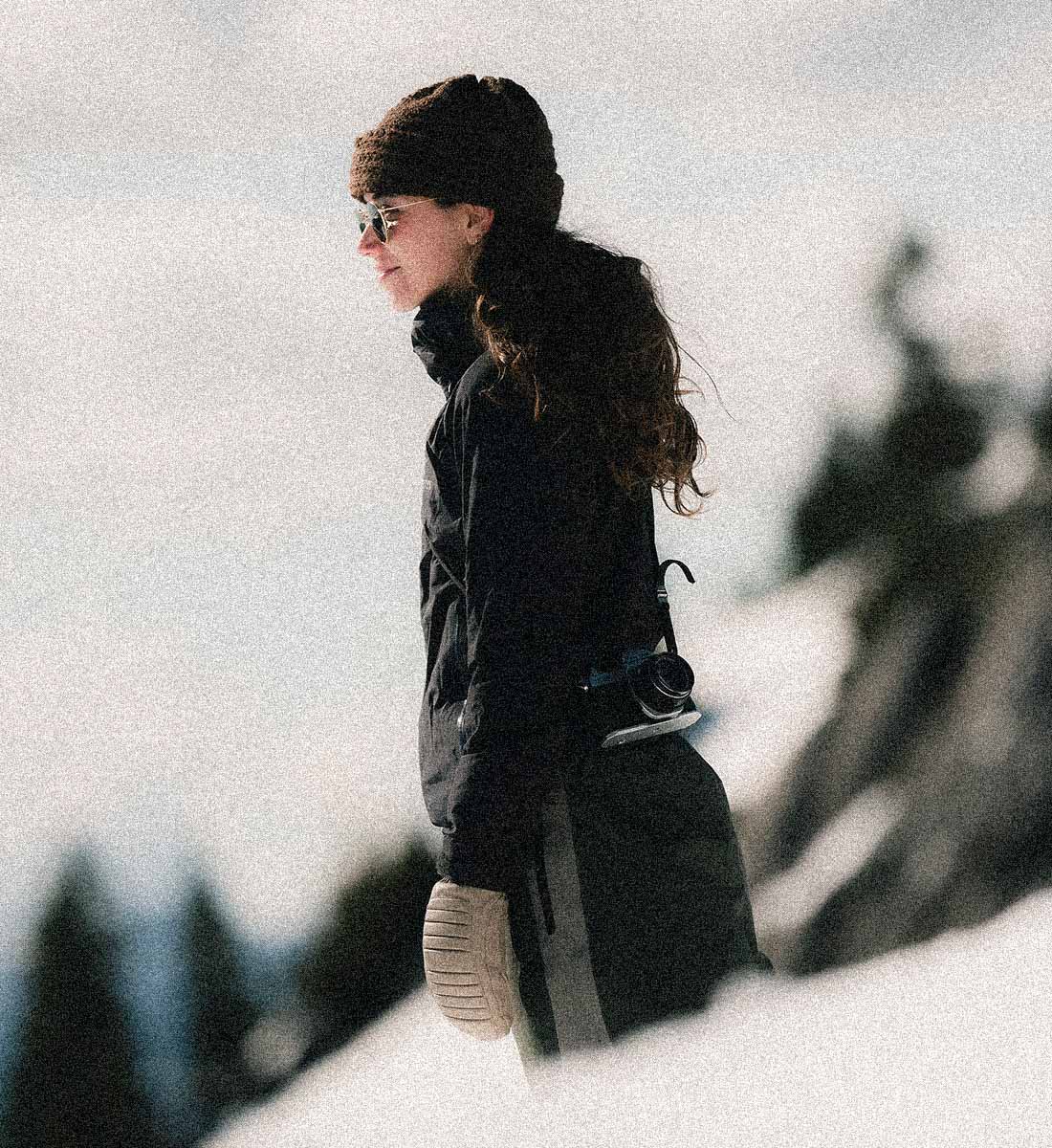 A woman in a black winter jacket, matching beanie, and knit mitts stands in a snowy outdoor setting, looking off-camera. The background is heavily blurred with a grainy, stylized filter.