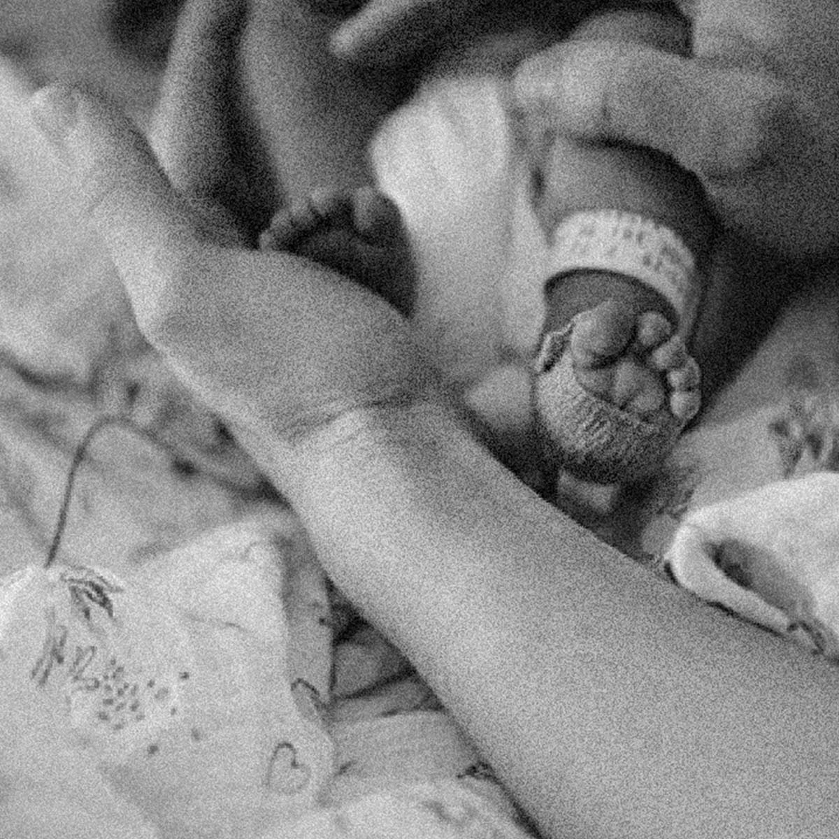 Black and white close-up photograph of a newborn baby's bare feet and toes, one wrapped in a knit bootie, resting on a mother's hand and held in another hand, with a hospital wristband visible.