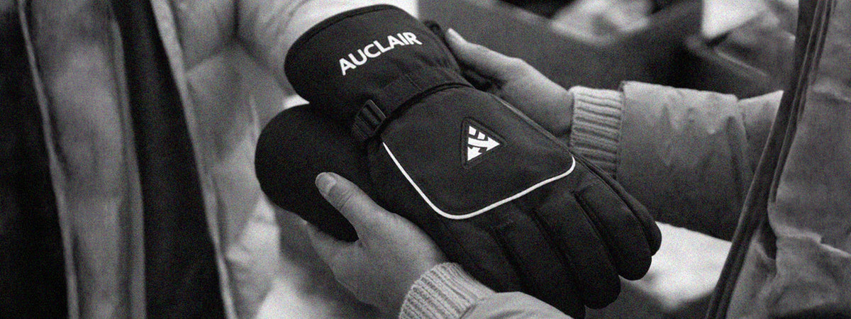 Black insulated winter gloves with white piping and branding, being worn by a person in a close-up black and white photo.