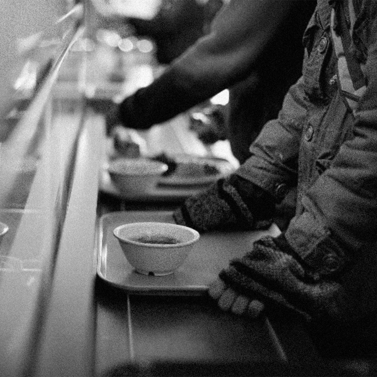 Black and white photograph of a person in a heavy jacket and knit gloves holding a cafeteria tray with a small bowl, with another tray and person blurred in the background.