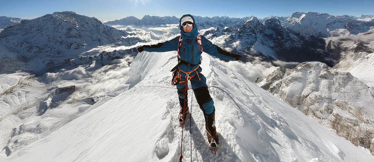 Person on a snowy mountain peak with arms outstretched, wearing climbing gear.