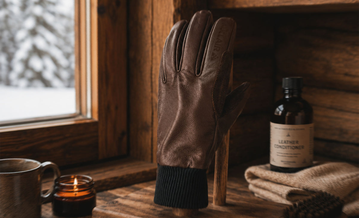 A brown leather winter glove with a black ribbed cuff stands upright on a wooden surface in a rustic cabin setting. A bottle labeled 'LEATHER CONDITIONER' and a folded tan cloth sit nearby, with a window showing snow outside and a candle on the left.