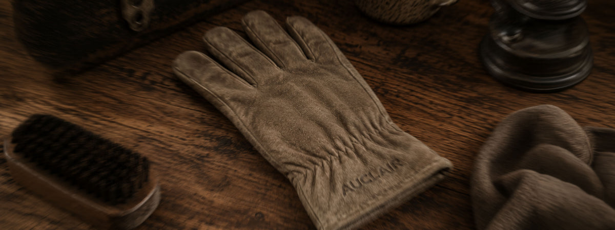 A single beige leather glove lies on a dark wooden surface, surrounded by rustic props including a brush, a folded matching cloth, and a lantern. The glove has 'MCGREGOR' embroidered near the cuff.