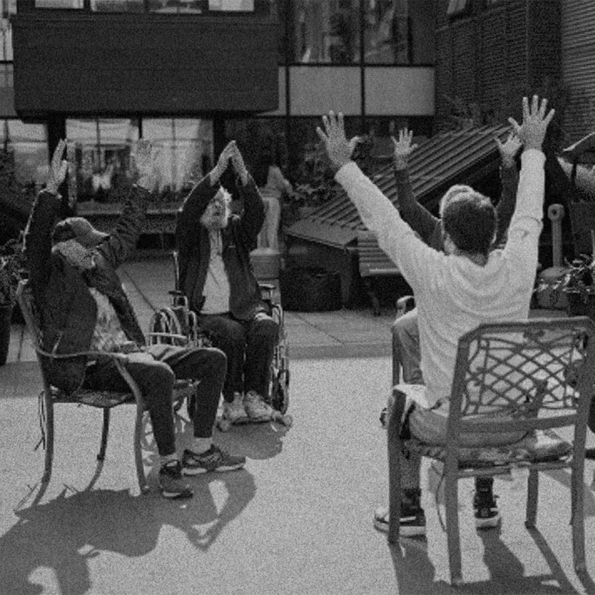 Black and white photograph of three people sitting in outdoor metal chairs in a courtyard, raising their hands in a synchronized gesture. The image has a high-contrast, grainy aesthetic.