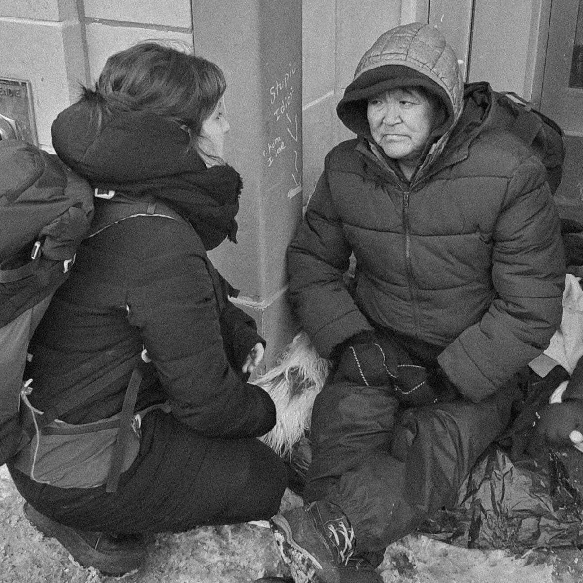 Black and white photograph showing two people in heavy winter clothing sitting or crouching near a building corner, with one person facing the other.