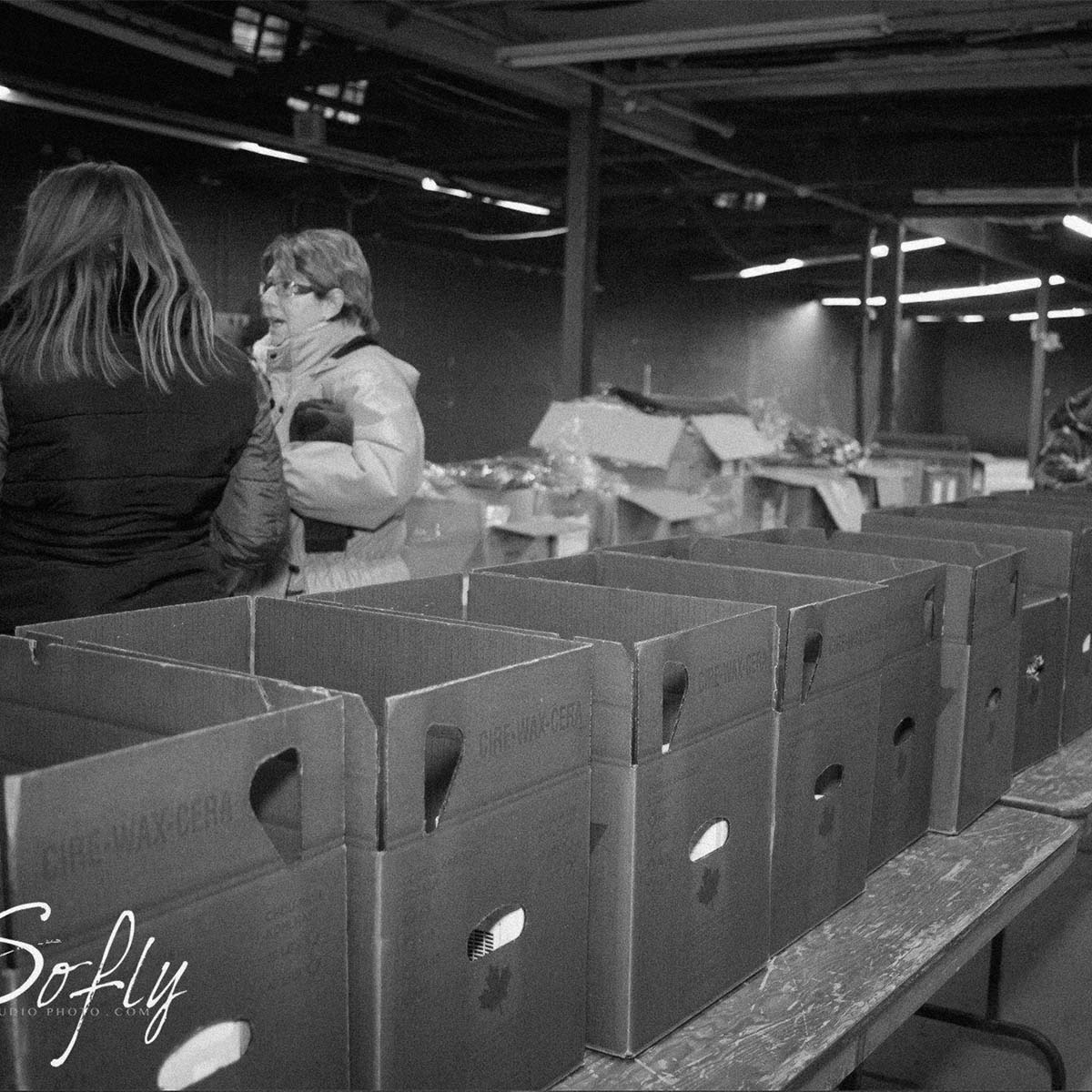 A black and white photograph showing two women standing in a warehouse or storage area next to several cardboard boxes on a table. The image has a visible 'Sofly' watermark in the lower-left corner.