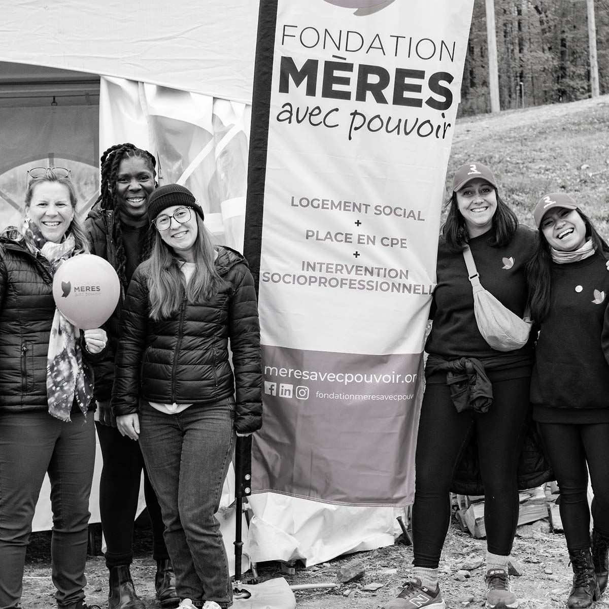 Five women stand together outdoors in front of a large vertical banner reading 'FONDATION MÈRES avec pouvoir' in black text on a white background. The photo is monochrome, and the women are wearing dark jackets, pants, and hats.