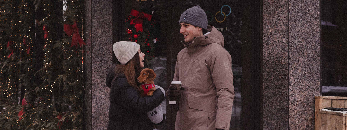 A man and a woman wearing winter clothing stand outside a building near a window with holiday decorations. The man wears a brownish-beige winter jacket, gray beanie, and dark gloves, while the woman wears a black coat, white beanie, and holds a small dog.