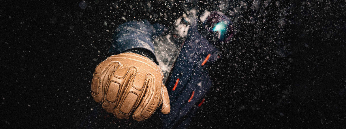 A close-up of a tan leather glove with a textured, padded design, shown against a dark, snowy background with a blurred figure in a blue jacket.