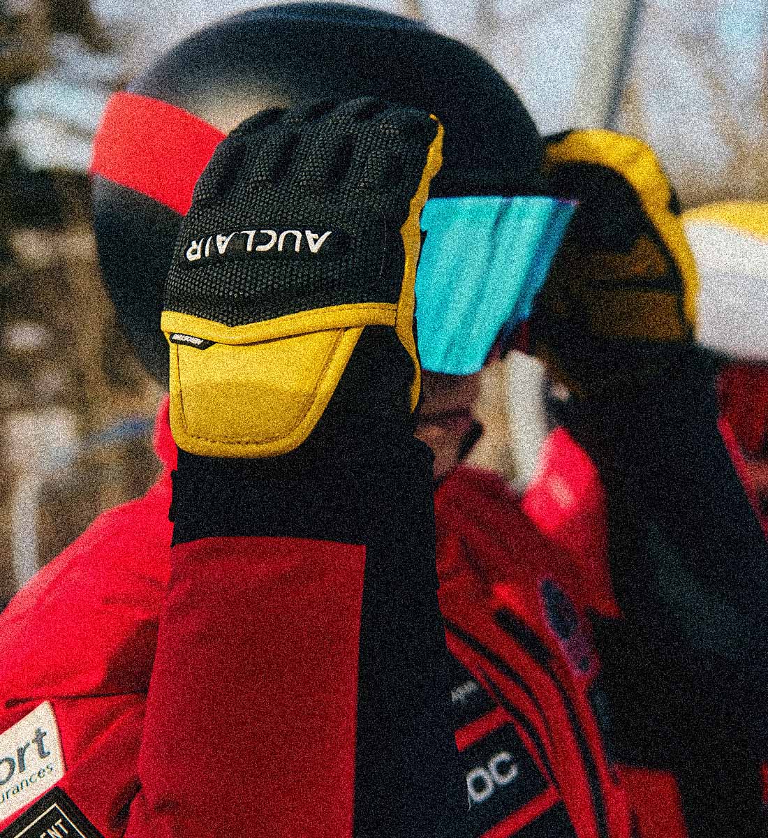 A close-up of a person wearing a black helmet with a bright blue visor and black and yellow gloves. The glove has the brand name 'AUKLAIR' printed in white on the back.
