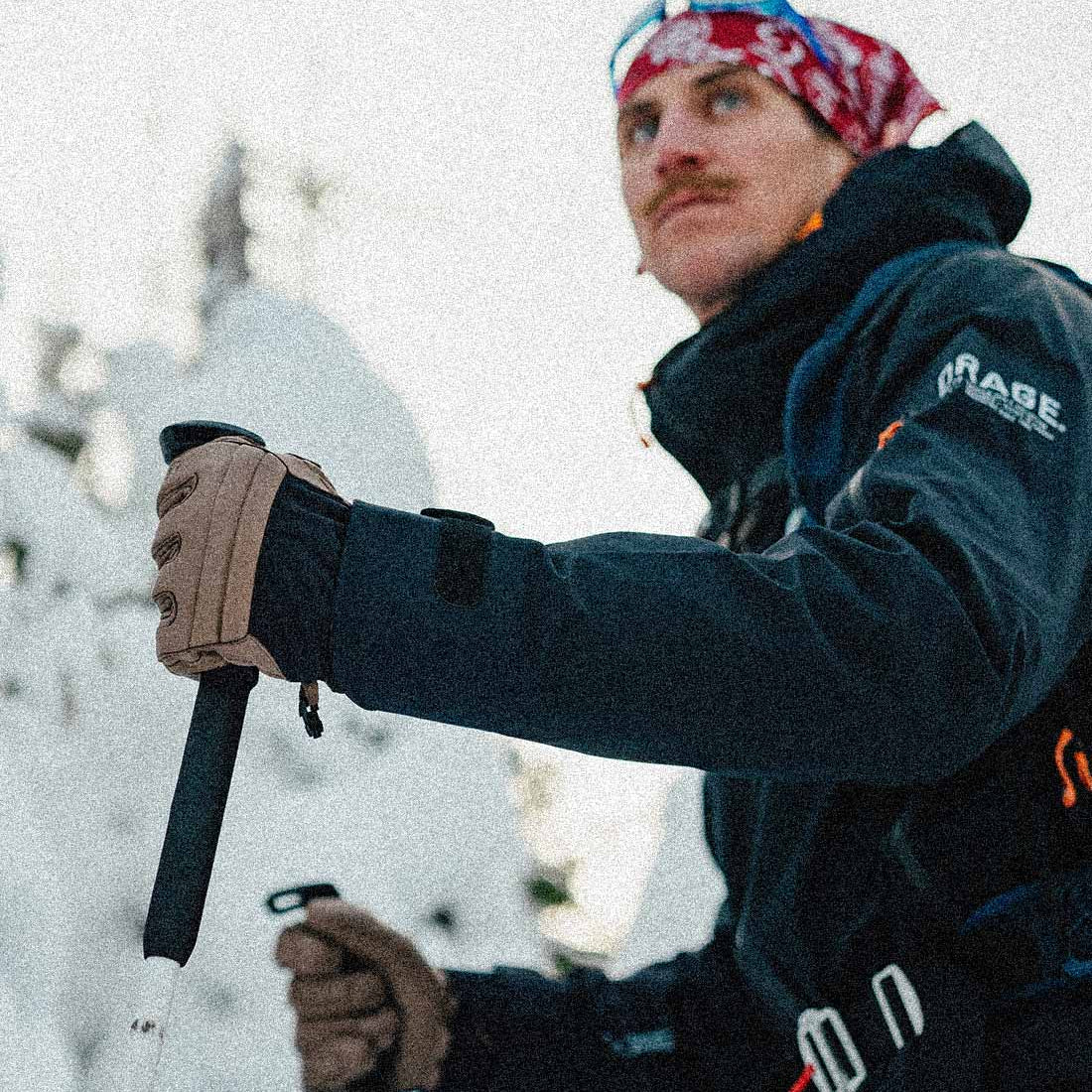 A man wearing a red, white, and blue patterned bandana, a dark ski jacket with 'KRABE' on the sleeve, and tan gloves holds a ski pole in a snowy outdoor setting.