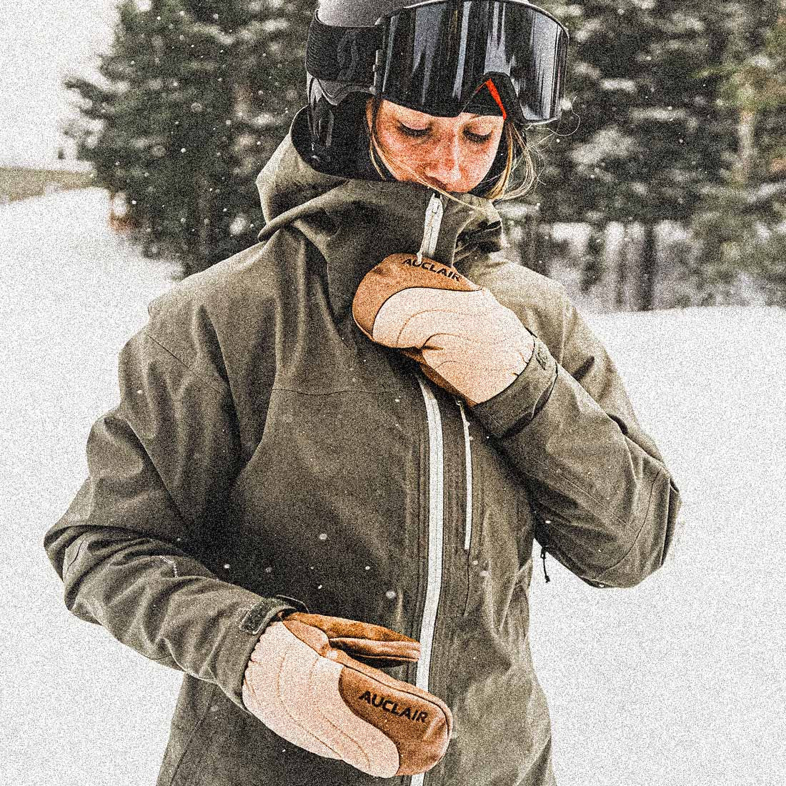 A person in a snowy outdoor setting wearing an olive green ski jacket, a dark helmet with goggles, and tan winter gloves with the brand name 'AUKLAIR' visible on the back of one glove.