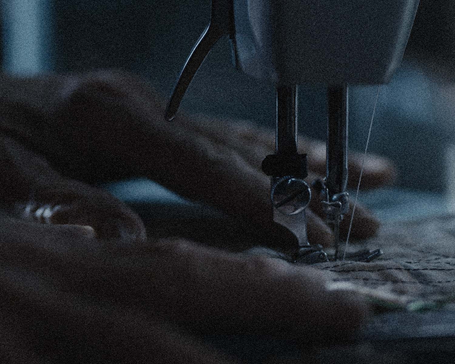 A close-up, low-light shot of a person's hands guiding fabric under the presser foot of a sewing machine.