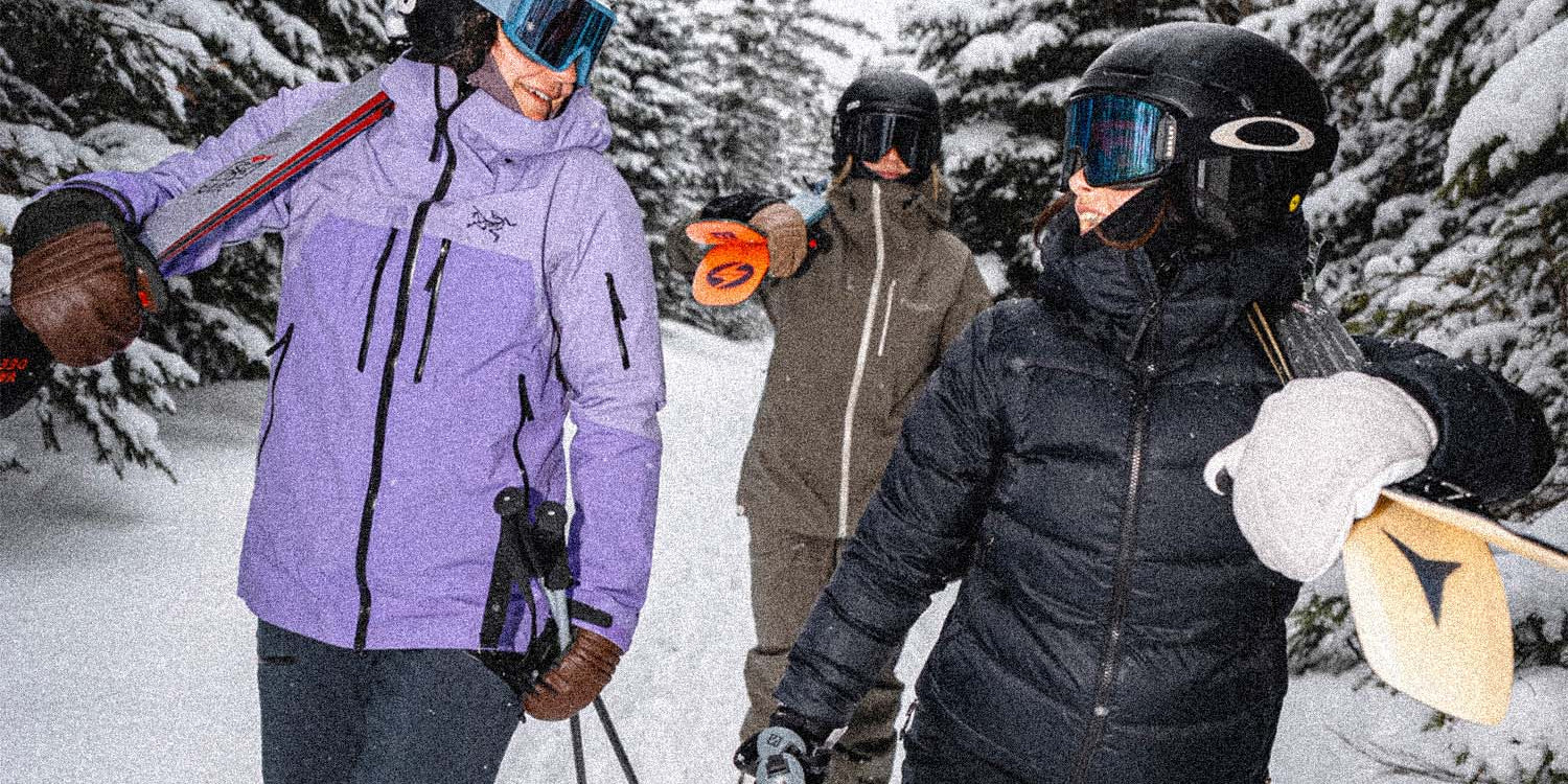 Three skiers walking through deep snow in a pine forest, wearing helmets, goggles, jackets, and ski pants. One skier in a light purple jacket carries skis over their shoulder, while the others carry ski poles.