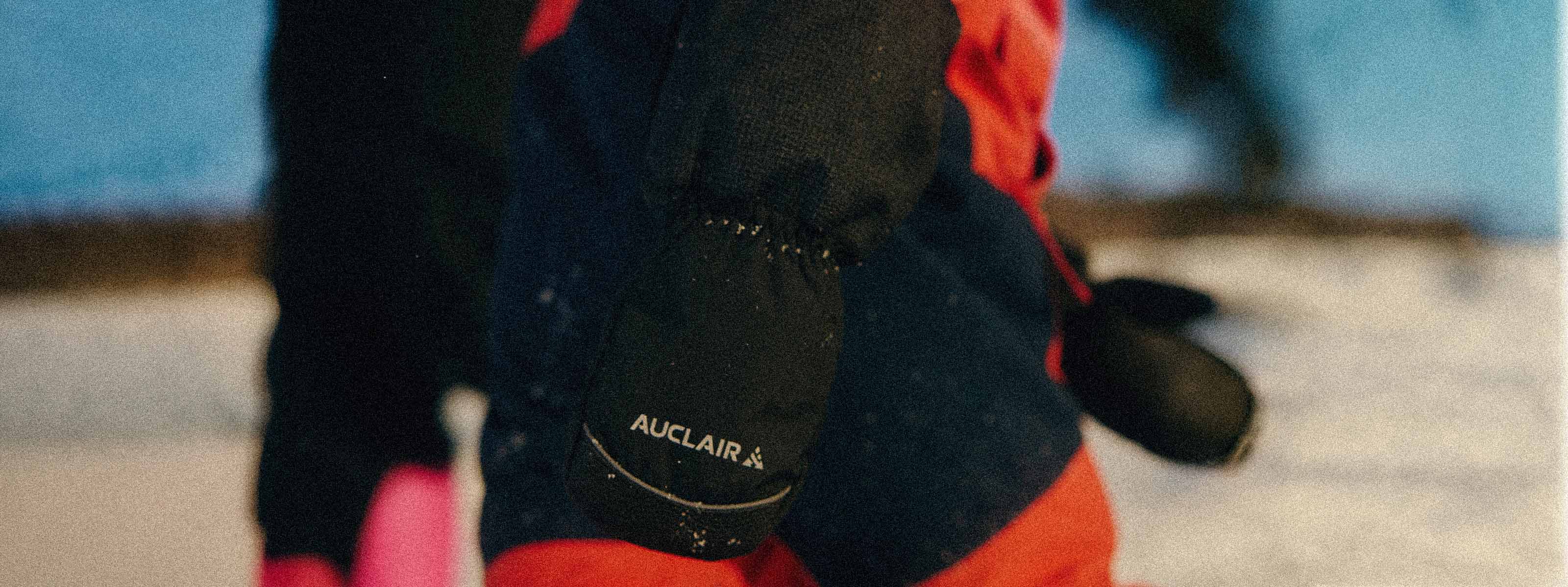 A close-up of a person wearing a black and red winter jacket, focusing on the sleeve with the brand name 'AUCLAIR' printed in white on the cuff, set against a snowy outdoor background.