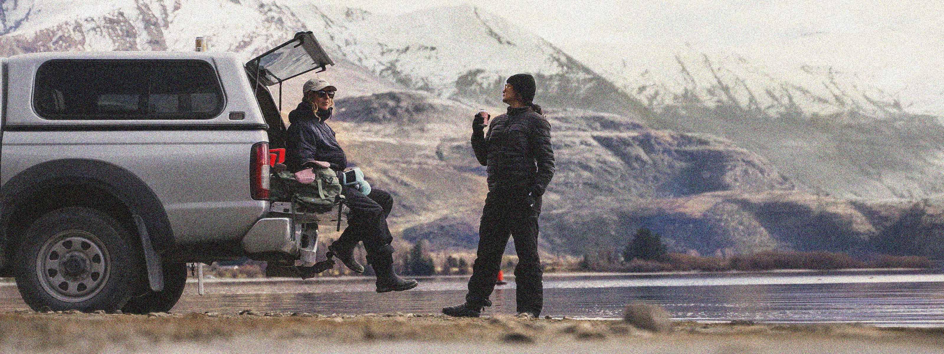A man in a black jacket stands near the open trunk of a silver pickup truck beside a lake, with another person sitting in the truck bed, set against a backdrop of snowy mountains and a river.